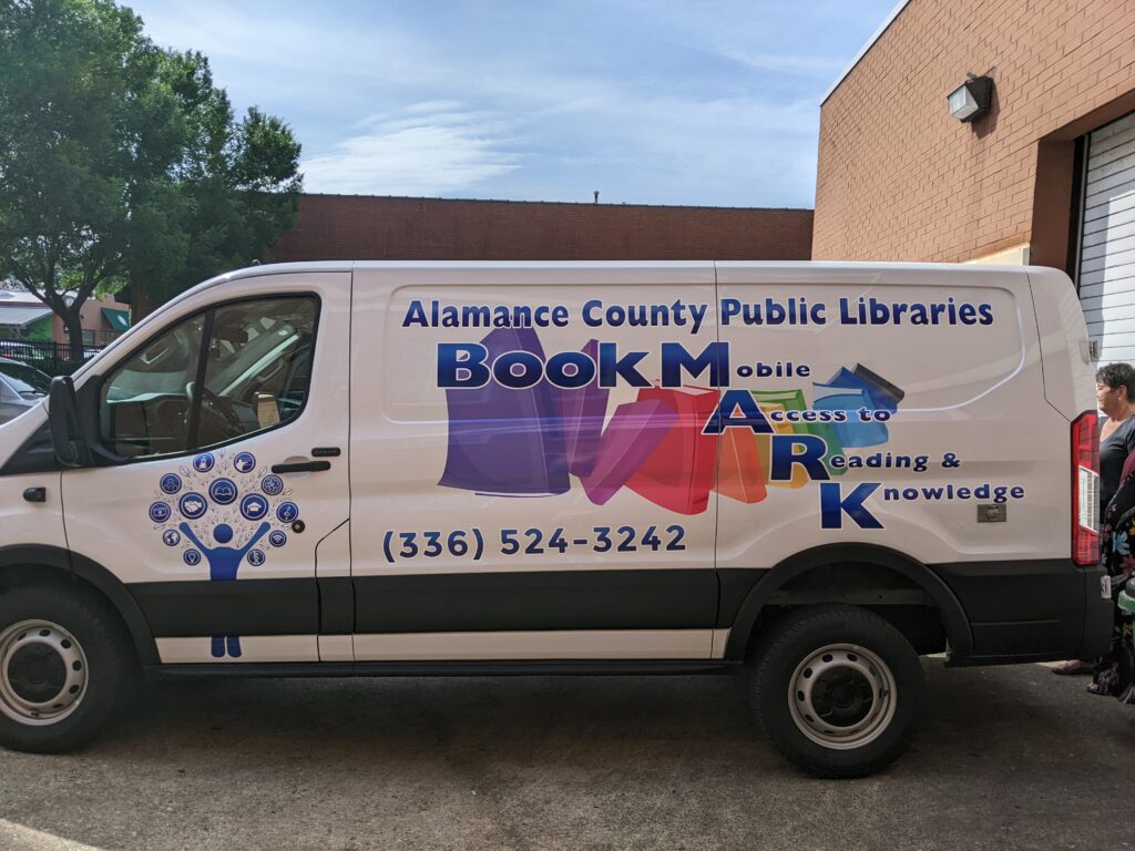Photo of BookMARK van. Side of van has the library logo on the door, and a colorful cascade of books, with the words "Alamance County Public Libraries BookMARK". Beside the last four letters it says, Mobile Access to Reading and Knowledge.