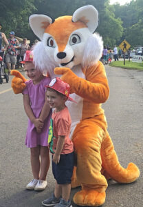 A person in an orange fox costume ("Swanson" - the Alamance Parks mascot) takes a photo with a young boy and girl at a park festival